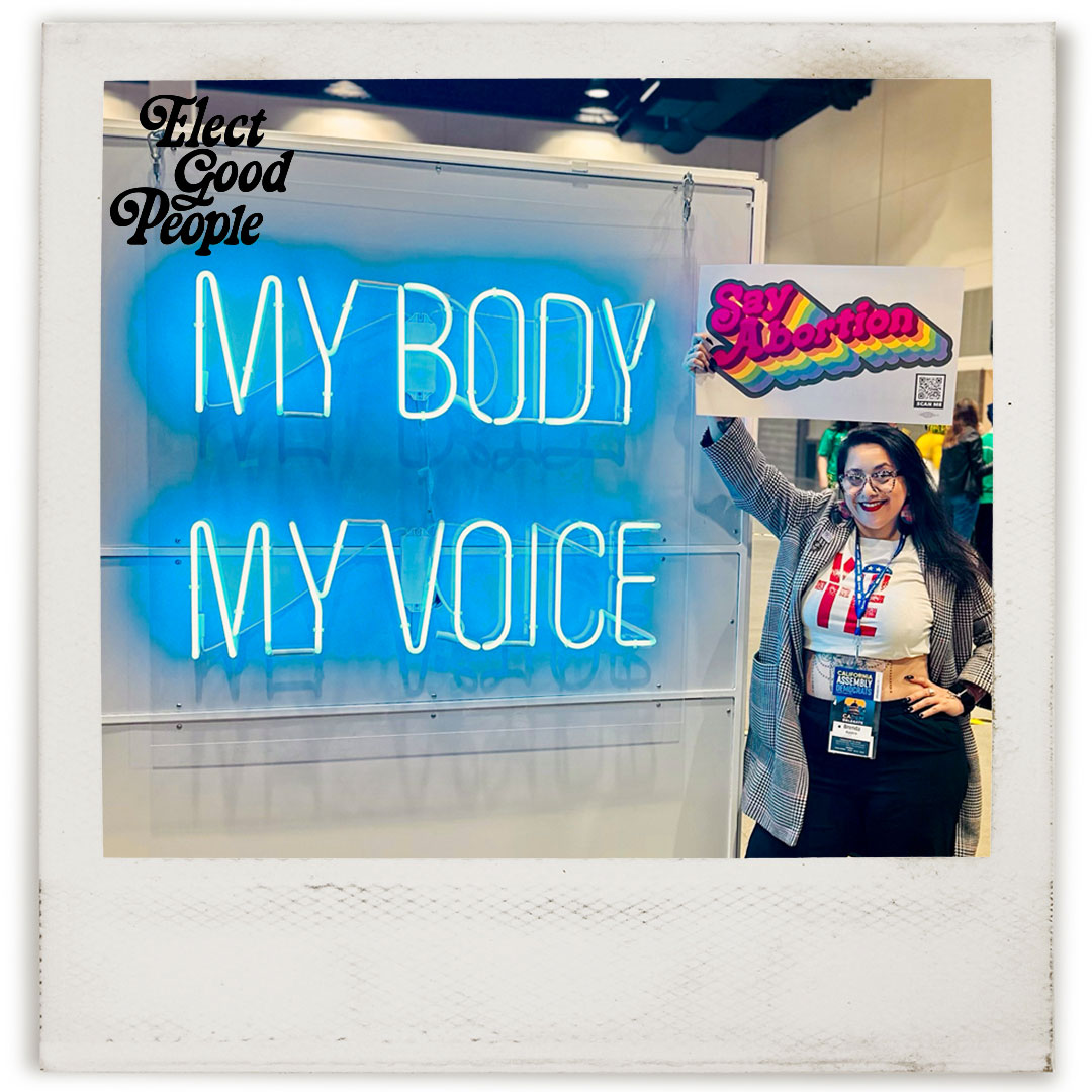 Candidate Brenda Aguirre posing in front of a sign that says "MY BODY MY VOICE"